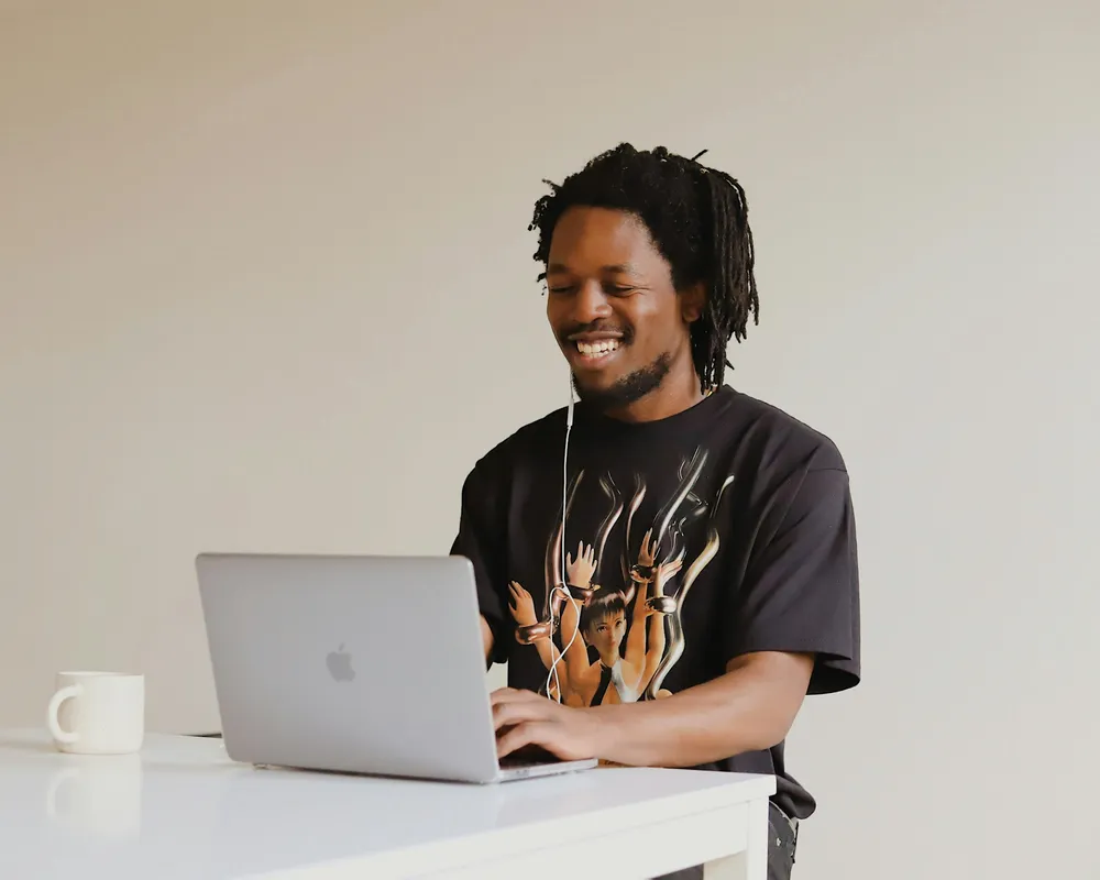 A man smiles while working on a laptop at a white desk.