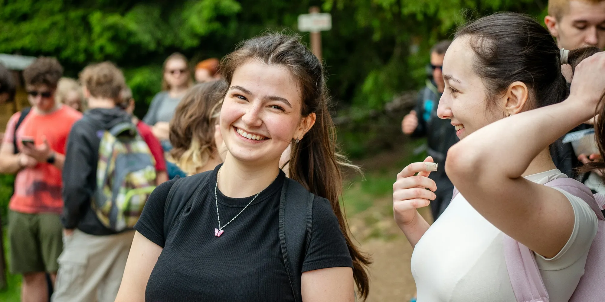A young woman smiles at a friend in a group of people with backpacks on a wooded trail.