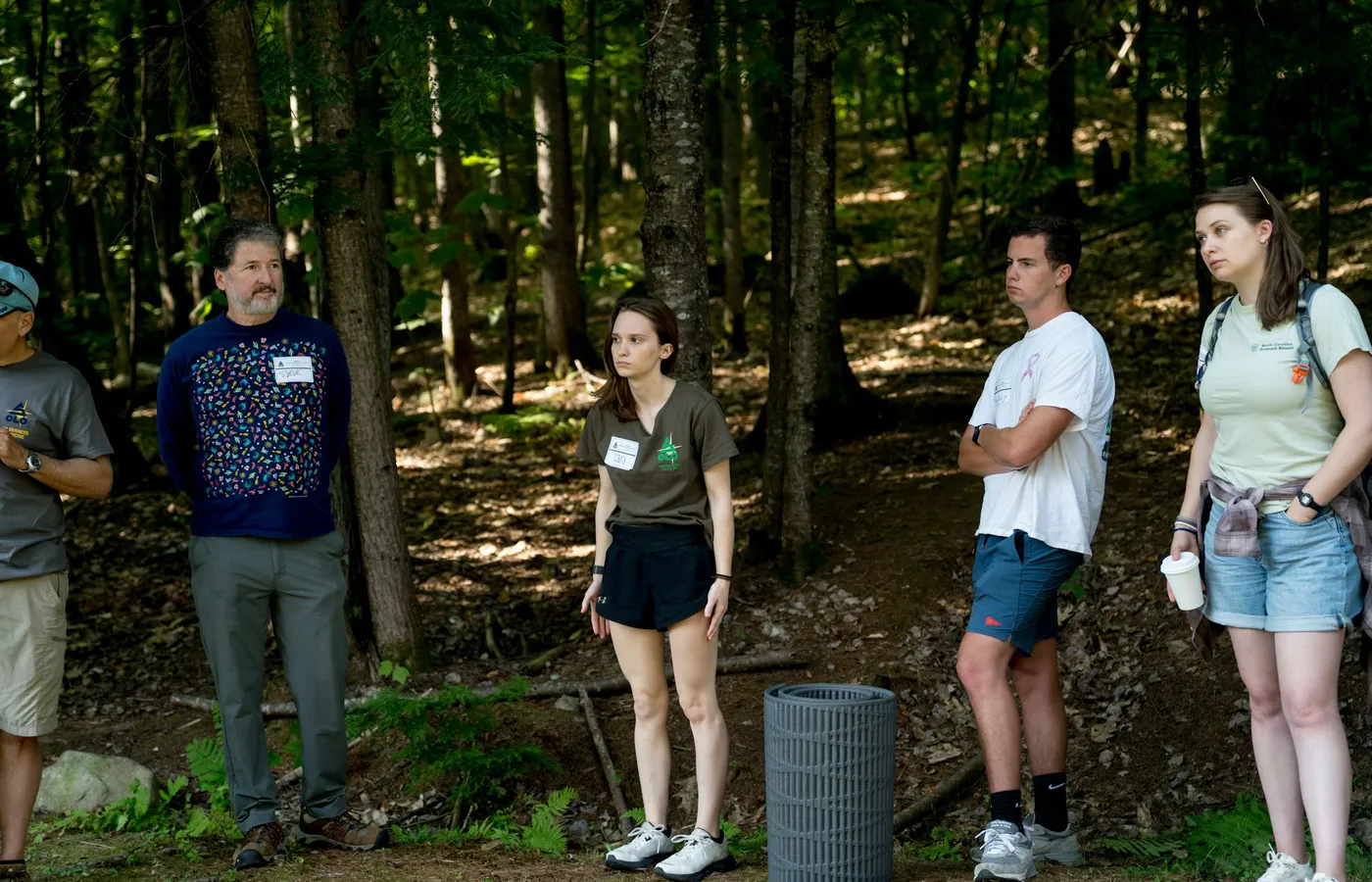 A small group of people stand together on a wooded path, some wearing name tags.