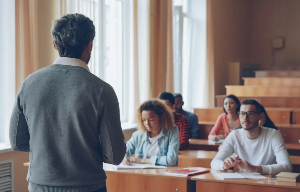 A professor speaks at the front of a lecture room while students take notes.