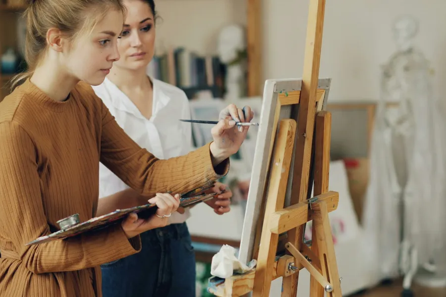 Two women stand at an easel in an art studio, one painting.