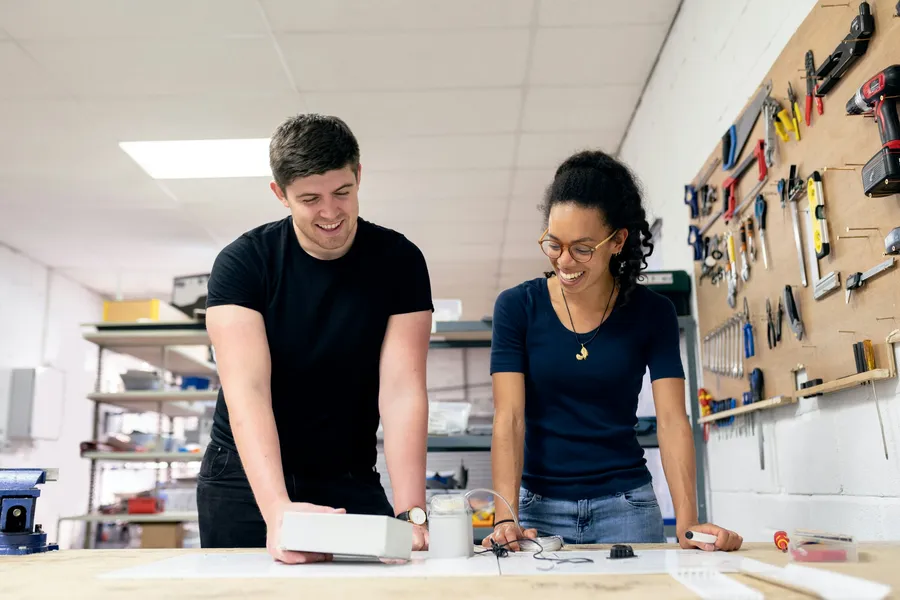 Two people smile over a workbench in a workshop with tools on the wall.