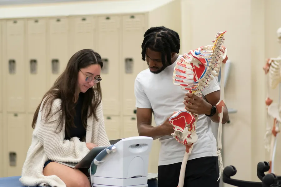 Two students in a classroom study an anatomical model of a spine.