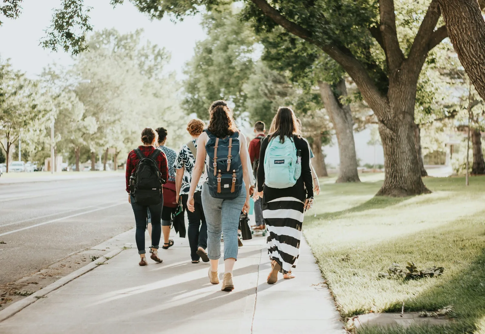 Students with backpacks walking down a tree-lined sidewalk on a sunny day.
