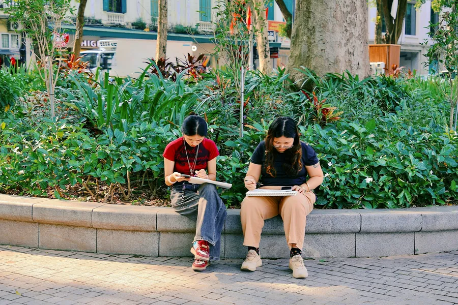 Two students sit on a low garden wall, drawing in sketchbooks.