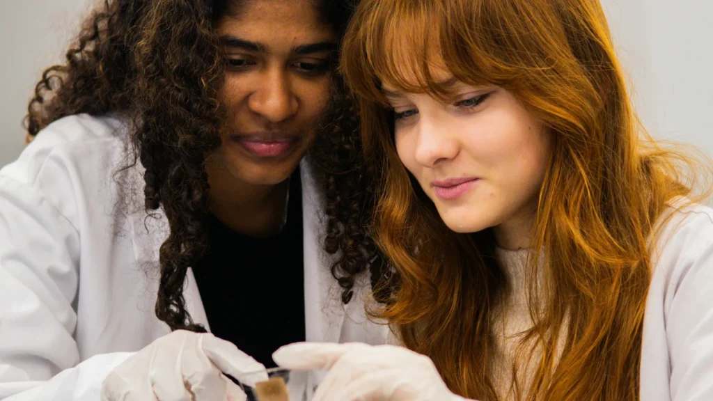 Two students in lab coats examine a small sample together.