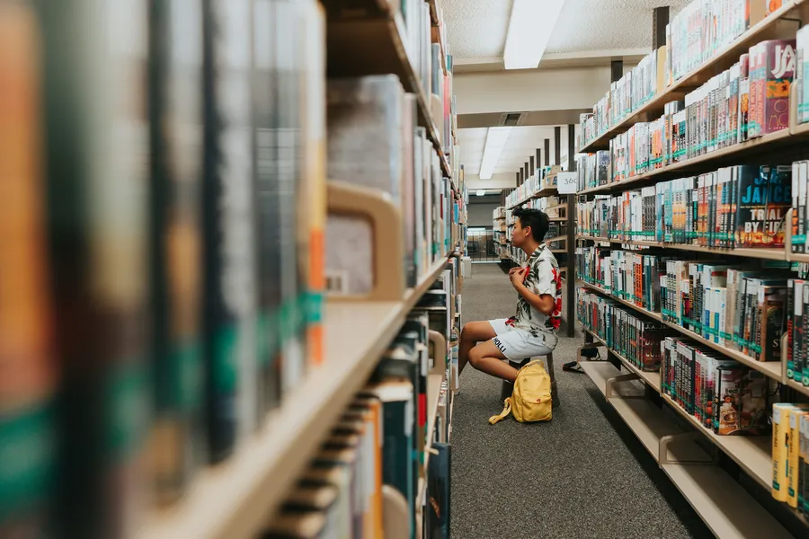A student sits on the floor between tall library shelves, reading.