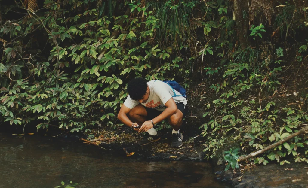 A young man crouches at the edge of a shaded stream, looking at something in his hands.