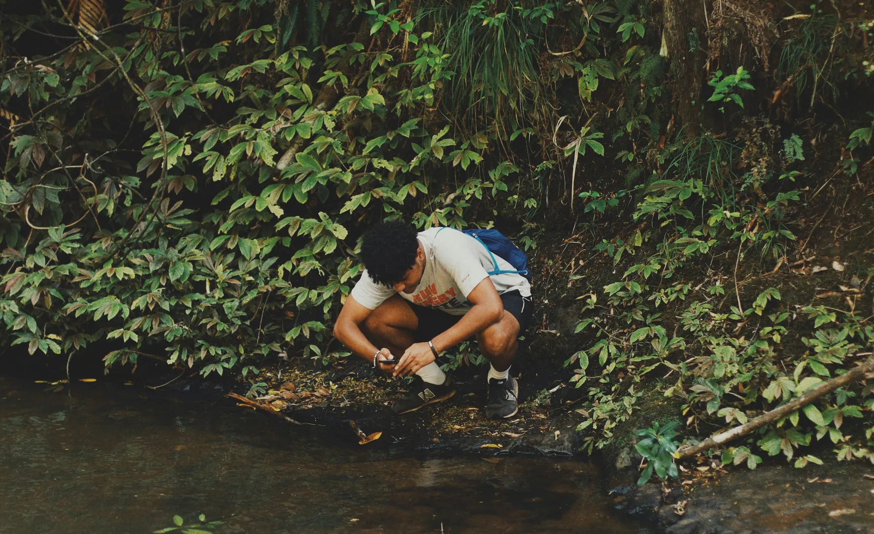 A young man crouches at the edge of a shaded stream, looking at something in his hands.